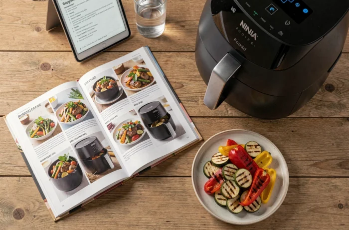 Livre de recettes Air Fryer Ninja Foodi ouvert sur une table en bois avec des légumes grillés et une tablette affichant un PDF
